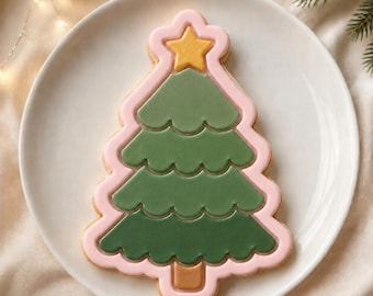 Puede incluir: Una galleta con forma de árbol de Navidad decorada con glaseado verde liso en tres secciones escalonadas, un borde rosa claro, una estrella amarilla en la parte superior y un tronco rectangular marrón, centrada en un plato de cerámica blanco.