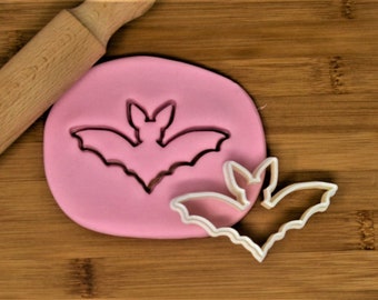 Könnte beinhalten: Ein weißer, plastikförmiger Fledermaus-Ausstecher auf einer Holzoberfläche mit rosa Modelliermasse. Der Ausstecher wird verwendet, um Fledermausformen aus der Masse auszuschneiden.