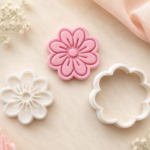 Peut inclure: Un ensemble de trois outils en forme de fleur comprenant un tampon à motif floral rose, un emporte-pièce blanc avec des détails de pétales internes et un emporte-pièce blanc à contour floral, tous avec un design à huit pétales arrondis.