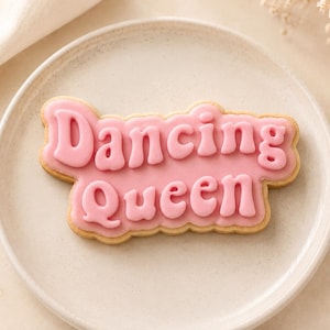 May include: A sugar cookie shaped like a speech bubble with light pink icing. The text Dancing Queen is written in a raised, darker pink, rounded font across the center of the cookie. The cookie is placed on a round, off-white ceramic plate.
