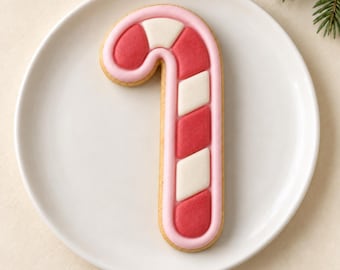 May include: A candy cane shaped cookie decorated with smooth light pink icing outlines and alternating diagonal segments of solid red and white icing. The cookie is centered on a plain white circular plate with a smooth, flat surface.