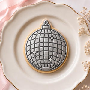 May include: A round sugar biscuit decorated with a silver-coloured icing design shaped like a disco ball. The icing features a grid pattern of square tiles and three four-pointed star accents, with a small loop at the top of the disco ball shape.