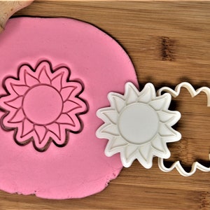 Puede incluir: Un cortador de galletas de girasol de plástico blanco sobre una superficie de madera con plastilina rosa. El cortador se está utilizando para cortar una forma de girasol de la plastilina.