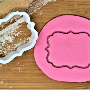Puede incluir: Un cortador de galletas de plástico blanco en forma de marco decorativo, con un recorte de fondant rosa sobre una superficie de madera.