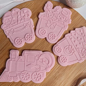 Puede incluir: Cuatro cortadores de galletas rosas con temática navideña sobre una superficie de madera. Los cortadores incluyen un tren, un árbol de Navidad y dos vagones diferentes con personajes festivos y golosinas. Los cortadores están detallados con ruedas y diseños festivos.