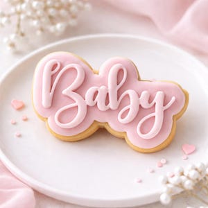 May include: A pink iced biscuit in a cloud shape, with the word "Baby" written in cursive. The biscuit sits on a white plate, with small pink heart-shaped sprinkles and decorative flowers in the background.