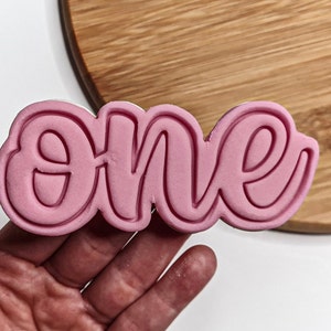 Puede incluir: Un cortador de galletas rosa con la palabra "one" en cursiva. El cortador de galletas se sostiene en una mano, con una tabla de cortar de madera en el fondo.