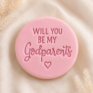 May include: A round, light pink biscuit featuring the embossed text "WILL YOU BE MY Godparents" in a combination of block and script lettering, with a small heart symbol centred directly below the text on the surface of the biscuit.