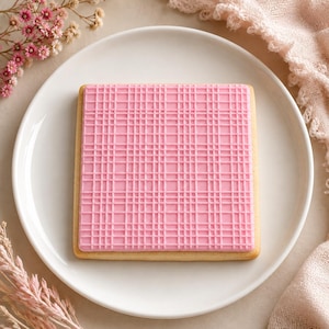 May include: A square sugar biscuit topped with a smooth layer of light pink icing featuring a raised, embossed grid pattern of intersecting horizontal and vertical lines. The biscuit is centred on a plain white plate, showcasing a clean, geometric design.