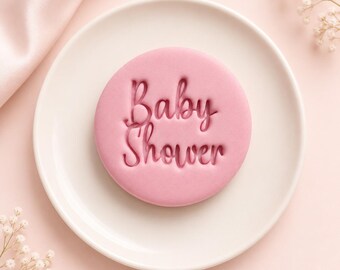 May include: A round, light pink biscuit featuring the embossed text Baby Shower in a cursive font. The biscuit is centred on a plain white plate, providing a clear view of the smooth surface and the indented lettering on the circular treat.