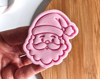 May include: A pink plastic Santa Claus face cookie cutter. The cutter features a detailed outline of Santa's hat, face, mustache, and beard. The cutter is held against a wooden surface.