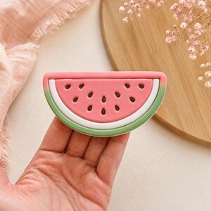 Peut inclure: Une barrette en forme de tranche de pastèque semi-circulaire avec un centre rose parsemé de pépins noirs en forme de goutte, une bordure blanche et un contour vert. L'accessoire présente une finition mate lisse et une forme courbée réaliste.