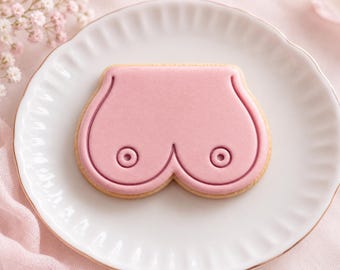 May include: A sugar cookie shaped like a pair of breasts, featuring smooth light pink icing with a raised outline and two small circular details representing nipples, centered on a white scalloped-edge ceramic plate.