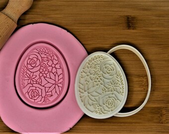 May include: A white plastic cookie cutter in the shape of an egg with a floral design. The cutter is on a wooden surface next to a pink silicone mold of the same design.