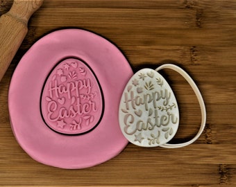 Puede incluir: Un molde de silicona rosa y un cortador de galletas de plástico blanco, ambos con forma de huevo con las palabras "Happy Easter" en su interior.