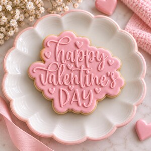 May include: A scalloped-edge sugar cookie with pink icing featuring the embossed text Happy Valentine's Day and small heart accents. The cookie is centered on a white scalloped plate with a light pink rim, resting on a marble surface.