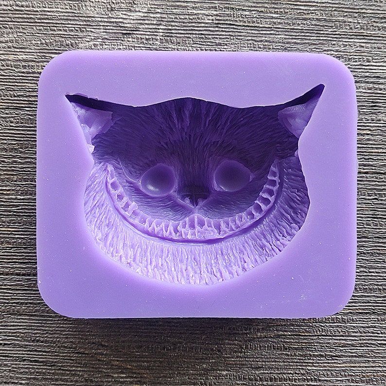 May include: A purple silicone mold featuring the face of the Cheshire Cat from Alice in Wonderland. The mold has a rectangular shape with rounded corners and a detailed depiction of the cat's grinning face, including its eyes, nose, and teeth.