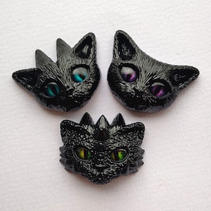May include: Three black cat head-shaped decorative items with iridescent eyes. One cat has teal eyes, another has purple eyes, and the third has green and yellow eyes. The cat heads have a textured, glossy finish.