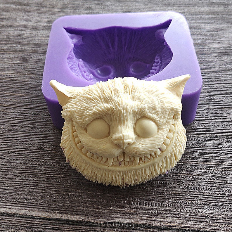 May include: A light beige Cheshire Cat sculpture with a wide grin, next to a purple silicone mold of the same cat face. The cat has large, empty eye sockets and detailed fur texture. The mold is a darker shade of purple.