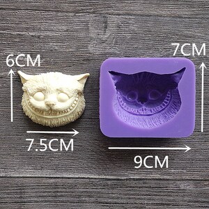 May include: A cream-colored Cheshire Cat face sculpture, 3 inches tall and 3 inches wide, next to a purple silicone mold of the same design, measuring 3.5 inches by 2.75 inches. The cat has a wide grin.