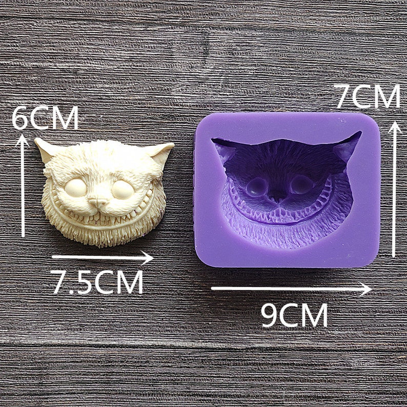 May include: A cream-colored Cheshire Cat face sculpture, 3 inches tall and 3 inches wide, next to a purple silicone mold of the same design, measuring 3.5 inches by 2.75 inches. The cat has a wide grin.