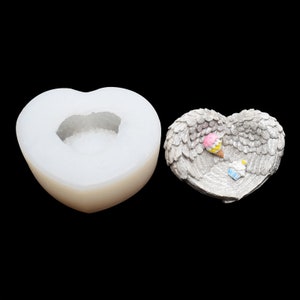 May include: Two heart-shaped items. One is a smooth, white mould with a central cavity. The other is a grey, heart-shaped dish with wing-like detailing. It has small, colourful ice cream and cupcake decorations.