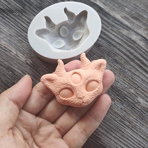 May include: A light peach-colored cat-faced mold and a finished cat face. The mold is white, and the cat face has three eyes and two horns. The cat face is held in a hand. The mold is made of silicone and is used for crafting.