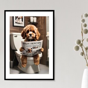 May include: A framed wall art print featuring a golden-brown puppy sitting on a white toilet while holding and reading a newspaper. The puppy has floppy ears and a dark collar. The background shows a bathroom setting with brown tiled walls.