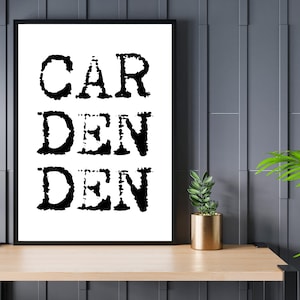 May include: Black and white typography print with the words "GARDEN DEN" on a white background. The print is framed in a black frame and is hanging on a gray wall.