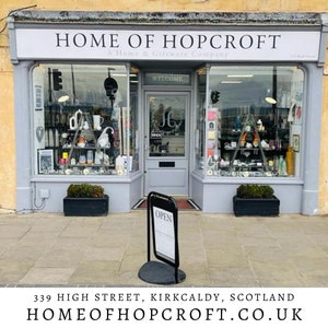 May include: A storefront with a gray facade and a sign that reads "HOME OF HOPCROFT A Home & Giftware Company". The storefront has two large windows and a glass door with the word "WELCOME" above it. There is a black sign on a stand in front of the door that reads "OPEN".