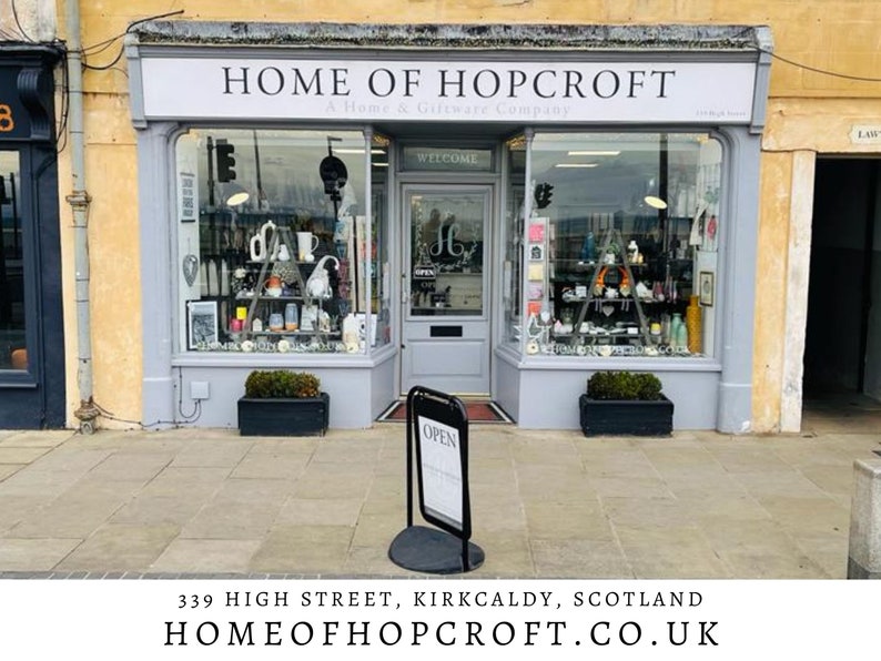 May include: A storefront with a gray facade and a sign that reads "HOME OF HOPCROFT A Home & Giftware Company". The storefront has two large windows and a glass door with the word "WELCOME" above it. There is a black sign on a stand in front of the door that reads "OPEN".