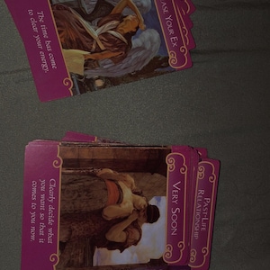 May include: A deck of rectangular oracle cards with magenta borders and gold filigree corners. The cards feature classical illustrations and text including Release Your Ex, Very Soon, and Past-Life Relationship. The text reads: The time has come to clear your energy. Clearly decide what you want so that it comes to you now. Release your exes and your soulmate will come very soon. They are a love from a past life.