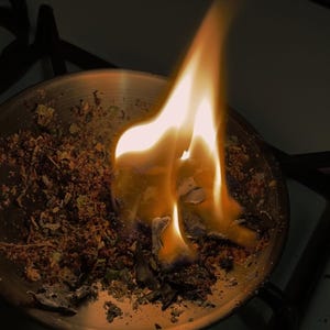 Peut inclure: Un bol rond en m&eacute;tal contenant un m&eacute;lange d'herbes s&eacute;ch&eacute;es et de mati&egrave;res botaniques qui br&ucirc;le activement avec une flamme orange et jaune vive. La flamme s'&eacute;l&egrave;ve du centre de la mati&egrave;re organique, projetant une lueur chaude sur le m&eacute;tal.