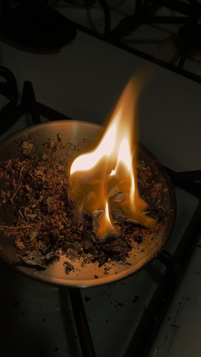 Peut inclure: Un bol rond en m&eacute;tal contenant un m&eacute;lange d'herbes s&eacute;ch&eacute;es et de mati&egrave;res botaniques qui br&ucirc;le activement avec une flamme orange et jaune vive. La flamme s'&eacute;l&egrave;ve du centre de la mati&egrave;re organique, projetant une lueur chaude sur le m&eacute;tal.