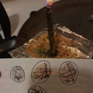 Peut inclure: Une bougie noire avec une flamme allum&eacute;e se trouve dans une po&ecirc;le en m&eacute;tal sur du papier aluminium recouvert d'herbes. Au premier plan, une bande de papier blanc pr&eacute;sente quatre symboles circulaires dessin&eacute;s &agrave; la main avec des lignes noires.