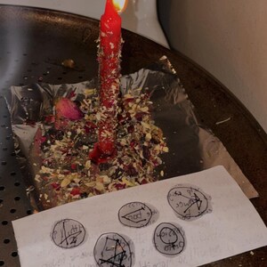 May include: A lit red candle covered in dried herbs and flower petals sits on a square of aluminium foil. In front of the candle, a white rectangular paper features five hand-drawn circular symbols with black ink lines and markings.