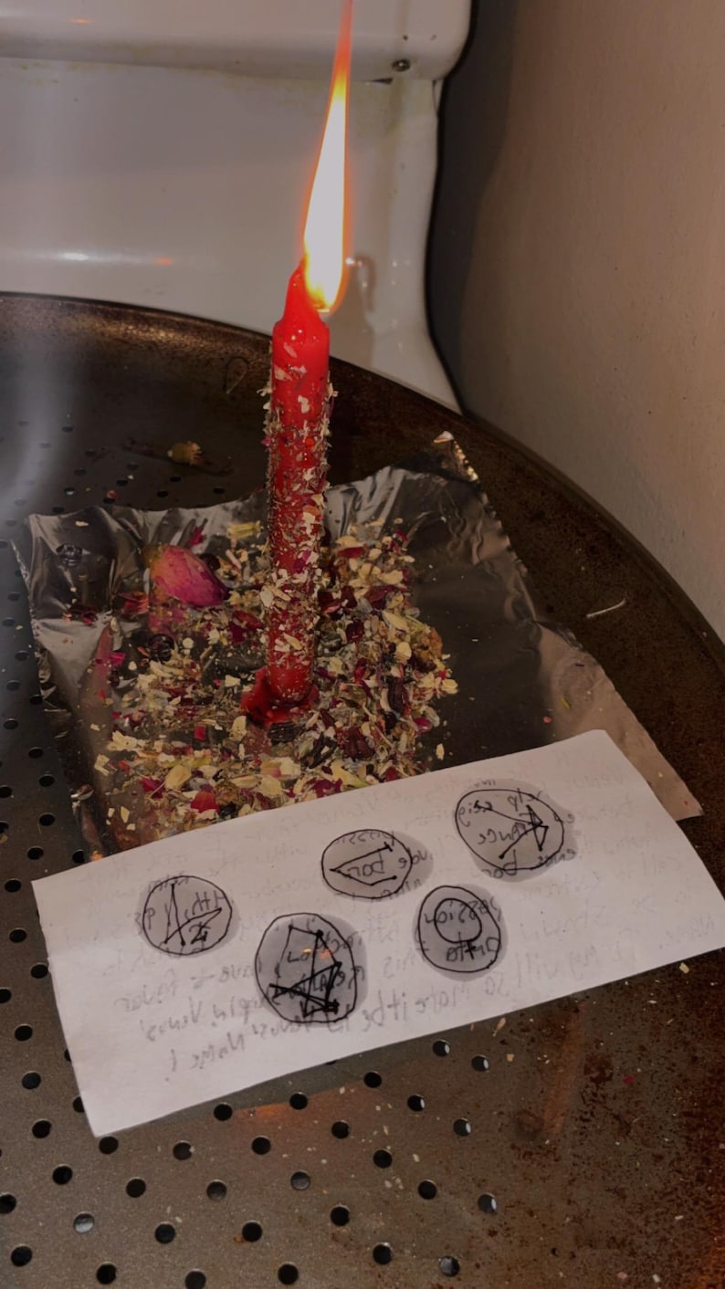 May include: A lit red candle covered in dried herbs and flower petals sits on a square of aluminium foil. In front of the candle, a white rectangular paper features five hand-drawn circular symbols with black ink lines and markings.