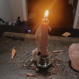 May include: A lit pink taper candle with melting wax dripping down its sides, held in a clear glass candle holder. The candle is surrounded by scattered dried herbs and small debris on a dark surface, with a large pink crystal nearby.
