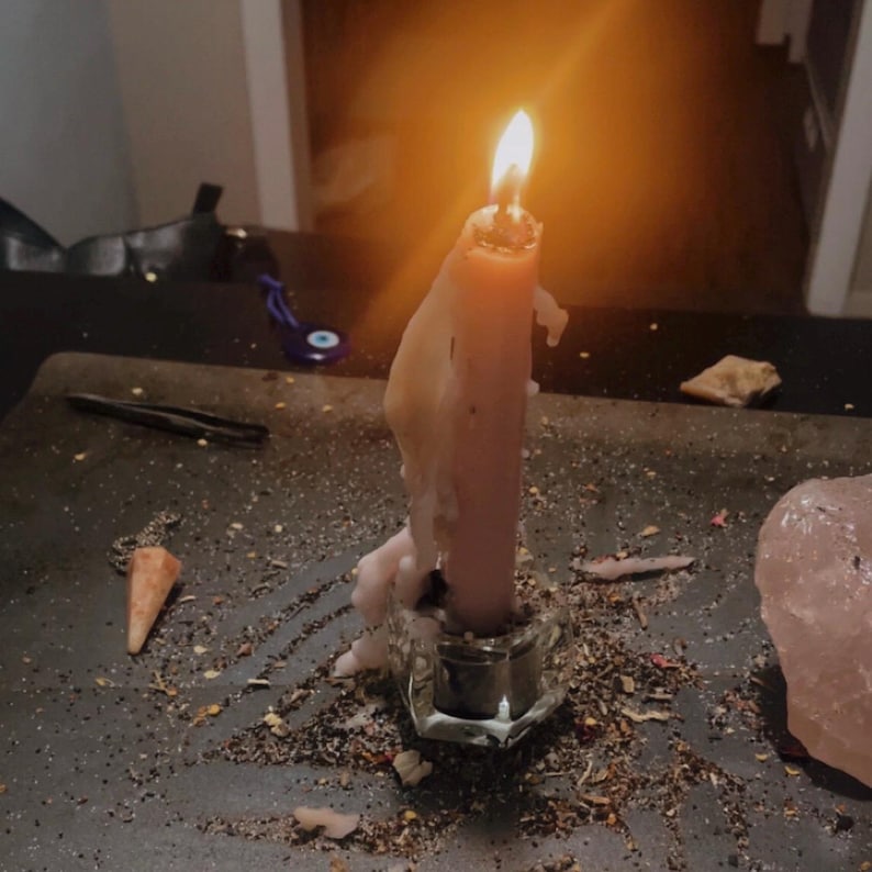 May include: A lit pink taper candle with melting wax dripping down its sides, held in a clear glass candle holder. The candle is surrounded by scattered dried herbs and small debris on a dark surface, with a large pink crystal nearby.
