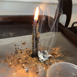Peut inclure: Une bougie noire allum&eacute;e est pos&eacute;e sur une feuille d'aluminium, entour&eacute;e de d&eacute;bris v&eacute;g&eacute;taux bruns et d'un petit morceau de papier blanc froiss&eacute; d&eacute;gageant de fines volutes de fum&eacute;e, avec un gobelet en plastique transparent contenant un liquide ambr&eacute;.