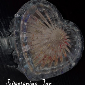 May include: A heart-shaped, clear glass jar with a textured, radial design. The jar contains colorful contents and the text "Sweetening Jar Group Spell". The jar sits on a dark surface.