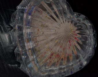 May include: A heart-shaped, clear glass jar with a textured, radial design. The jar contains colorful contents and the text "Sweetening Jar Group Spell". The jar sits on a dark surface.