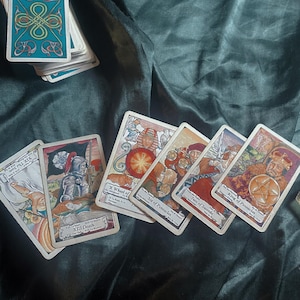 May include: A collection of tarot cards with detailed illustrations, including the Death card, the Wheel of Fortune, and the King of Pentacles. The cards are arranged on a dark green velvet surface, with a partial deck visible in the background.