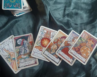 May include: Tarot cards featuring detailed illustrations, including the Death card and the Wheel of Fortune. The cards are displayed on a dark green, textured surface, with a small stack of cards visible in the background.