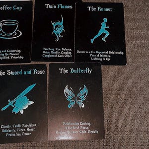 May include: Five rectangular oracle cards with black backgrounds and cyan blue illustrations and text. The cards feature titles and descriptions including Coffee Cup, Twin Flames, The Runner, The Sword and Rose, and The Butterfly, each with a corresponding icon.