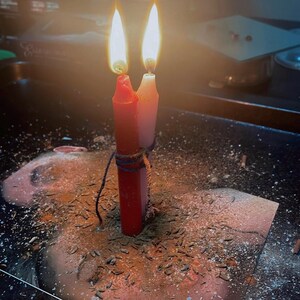 May include: Two cylindrical candles, one red and one white, are bound together with dark twine. Both candles are lit, producing bright flames. The base of the candles is surrounded by a scattering of small brown seeds and fine powder on a dark surface.