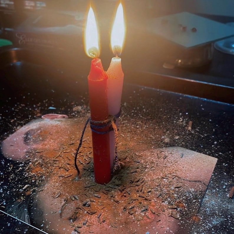 May include: Two cylindrical candles, one red and one white, are bound together with dark twine. Both candles are lit, producing bright flames. The base of the candles is surrounded by a scattering of small brown seeds and fine powder on a dark surface.