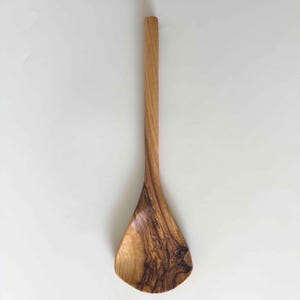May include: A wooden cooking spoon with a long, slender handle and a wide, rounded head. The spoon is made of light brown wood with a visible grain pattern. The spoon is designed for stirring and serving food.