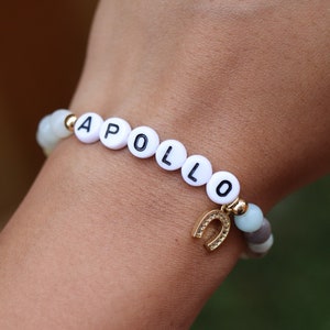 Könnte beinhalten: Ein weißes Perlenarmband mit dem Namen "APOLLO" in schwarzen Buchstaben. Ein goldener Hufeisen-Anhänger hängt am Armband.