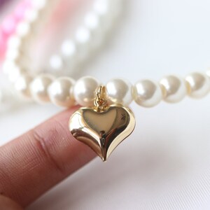 May include: A white pearl bracelet with a gold heart charm. The bracelet is on a finger.